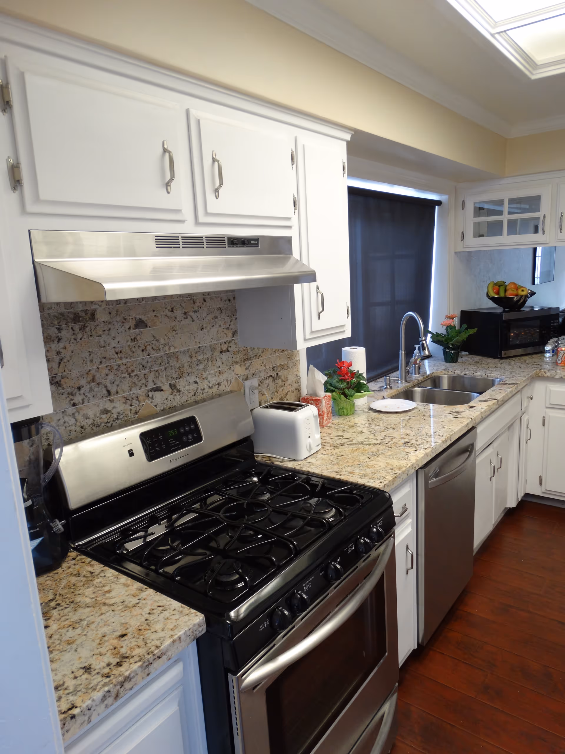 A modern kitchen with white cabinets, granite countertops, a stainless steel gas stove with oven, a stainless steel range hood, a toaster, a double sink, a dishwasher, a microwave, and a bowl of fruit on the counter. The floor is wooden, and there is a window with a dark roller shade.