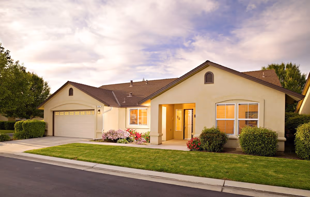 Single-story beige home with an attached garage, manicured front lawn, shrubs, and a lit entryway under a cloudy sky.