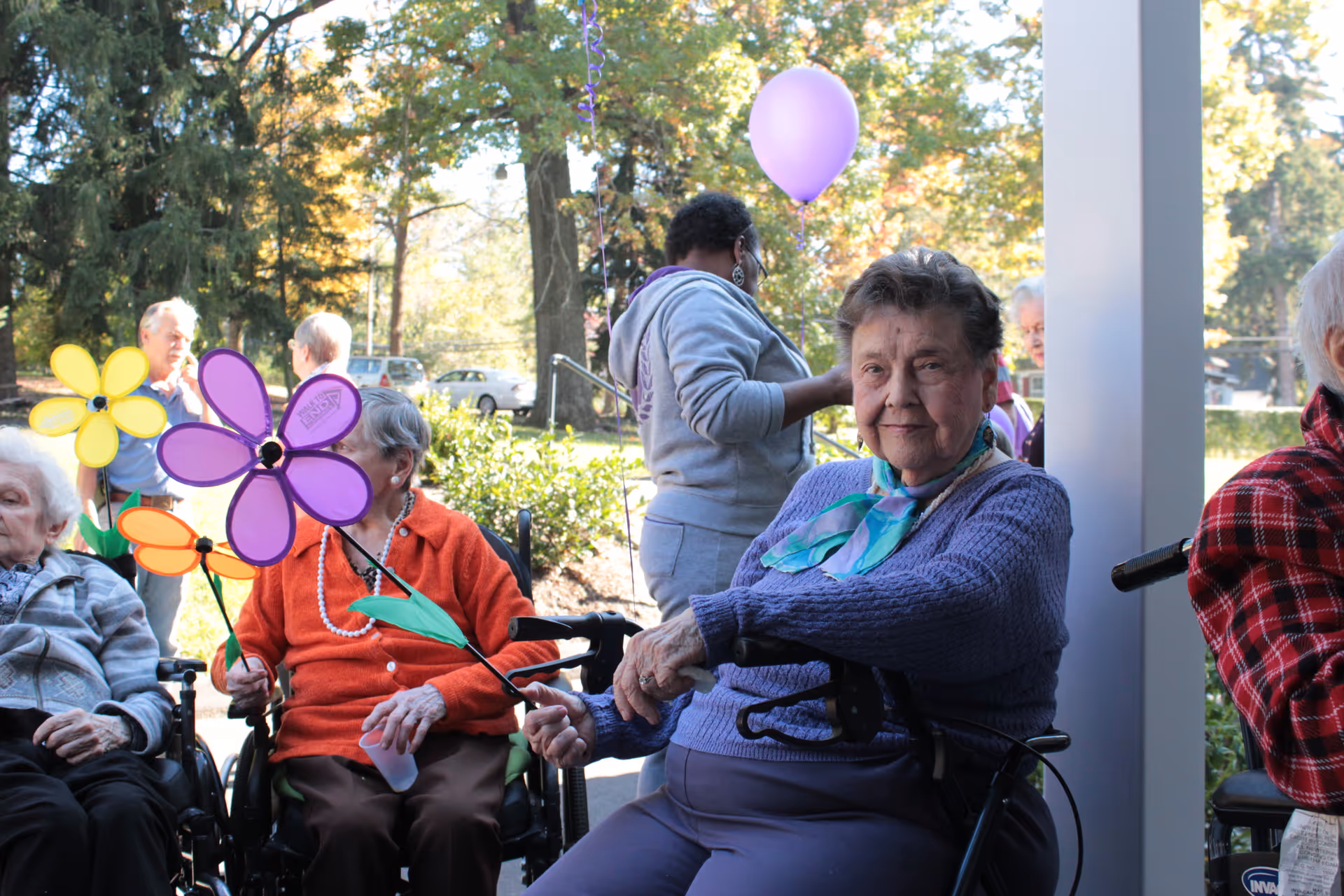 A group of elderly women sitting outdoors in wheelchairs, holding colorful flower-shaped pinwheels. One woman in a purple sweater and scarf smiles at the camera. In the background, a caregiver wearing a gray hoodie holds a purple balloon. Trees and parked cars are visible in the background.