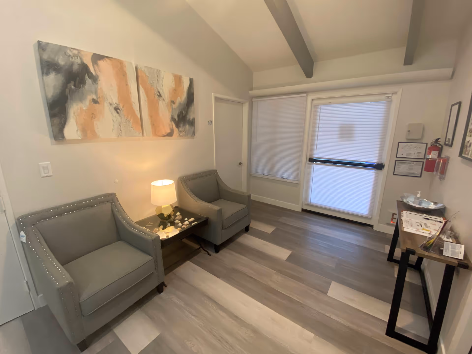 A small waiting area with two gray armchairs separated by a side table with a lamp and decorative shells. Above the chairs is a two-panel abstract painting in shades of gray, white, and peach. The room has light wood flooring, a door labeled with the number 10, and a window and door with closed blinds. A narrow console table on the right holds informational pamphlets, a hand sanitizer dispenser, and a fire extinguisher mounted on the wall above it.