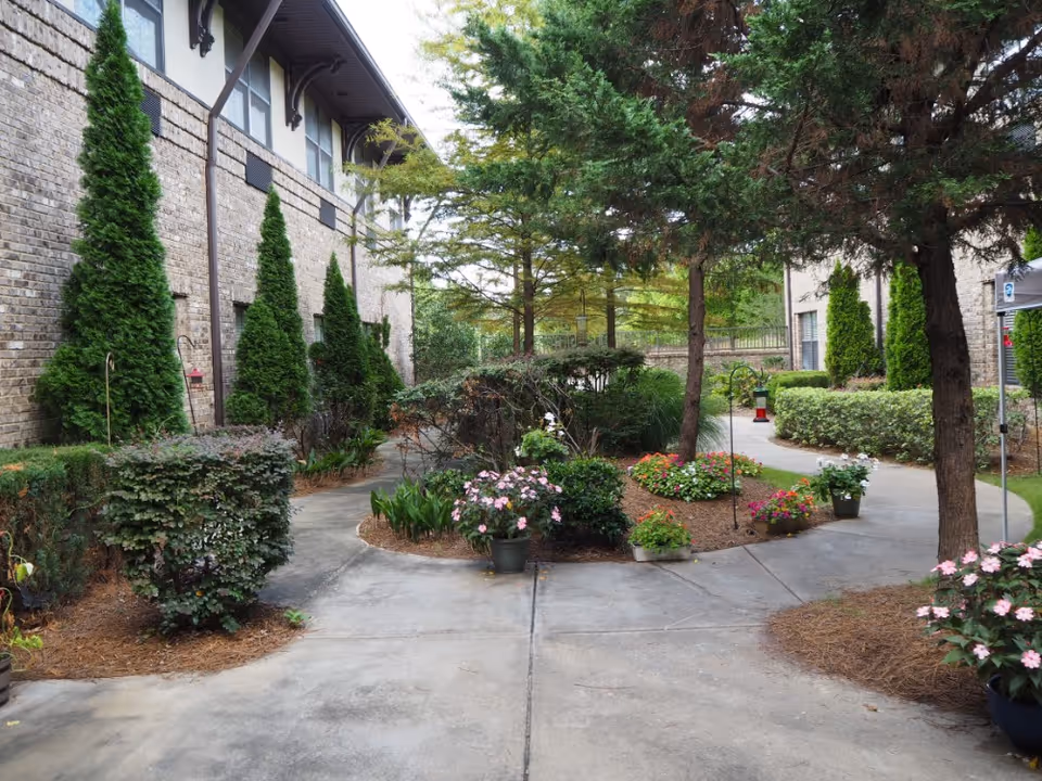 Outdoor garden area at Legacy Ridge Trussville with a paved walkway that splits into two paths surrounded by various green shrubs, trees, and colorful flowering plants. The building's brick exterior with windows and tall evergreen bushes lines one side of the walkway.