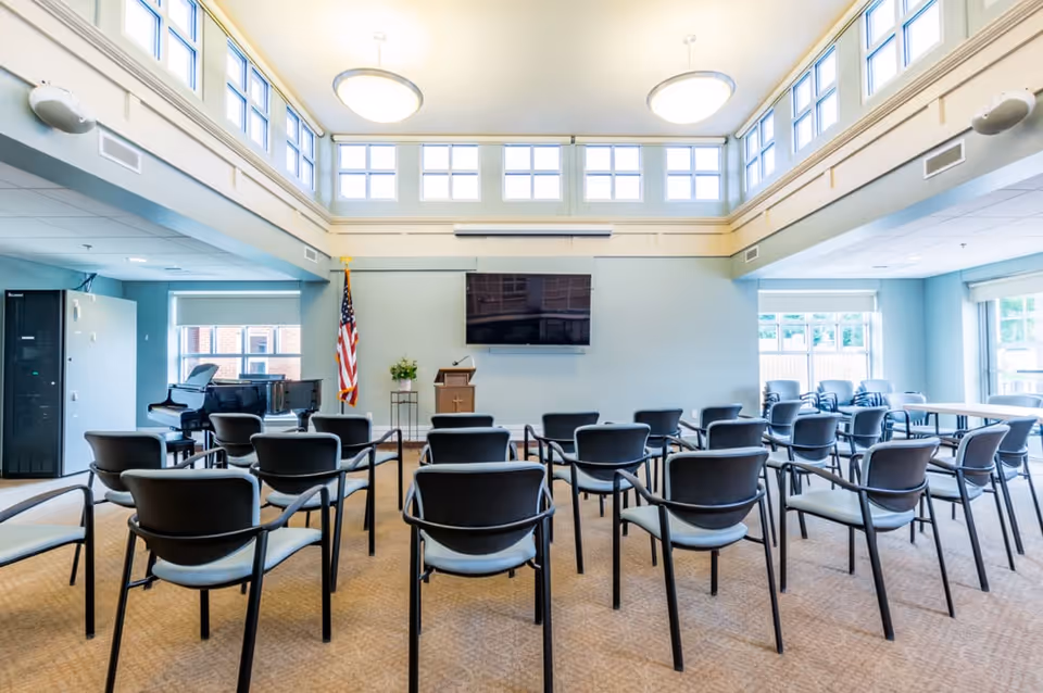 A bright meeting or activity room with rows of black chairs facing a wall-mounted flat screen TV and a wooden podium with a microphone. The room has high windows near the ceiling allowing natural light, an American flag, a piano in the corner, and light blue walls.