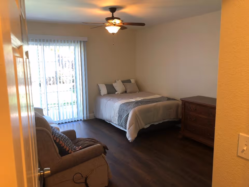 A bedroom with a bed made up with pillows and a comforter, a wooden dresser, a cushioned armchair with a patterned pillow, a ceiling fan with a light, and vertical blinds covering a sliding glass door that leads outside.