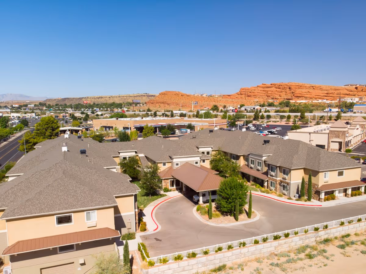 Aerial view of Spring Gardens St. George senior living facility with beige buildings, a covered entrance, surrounding parking lots, and red rock formations in the background under a clear blue sky.