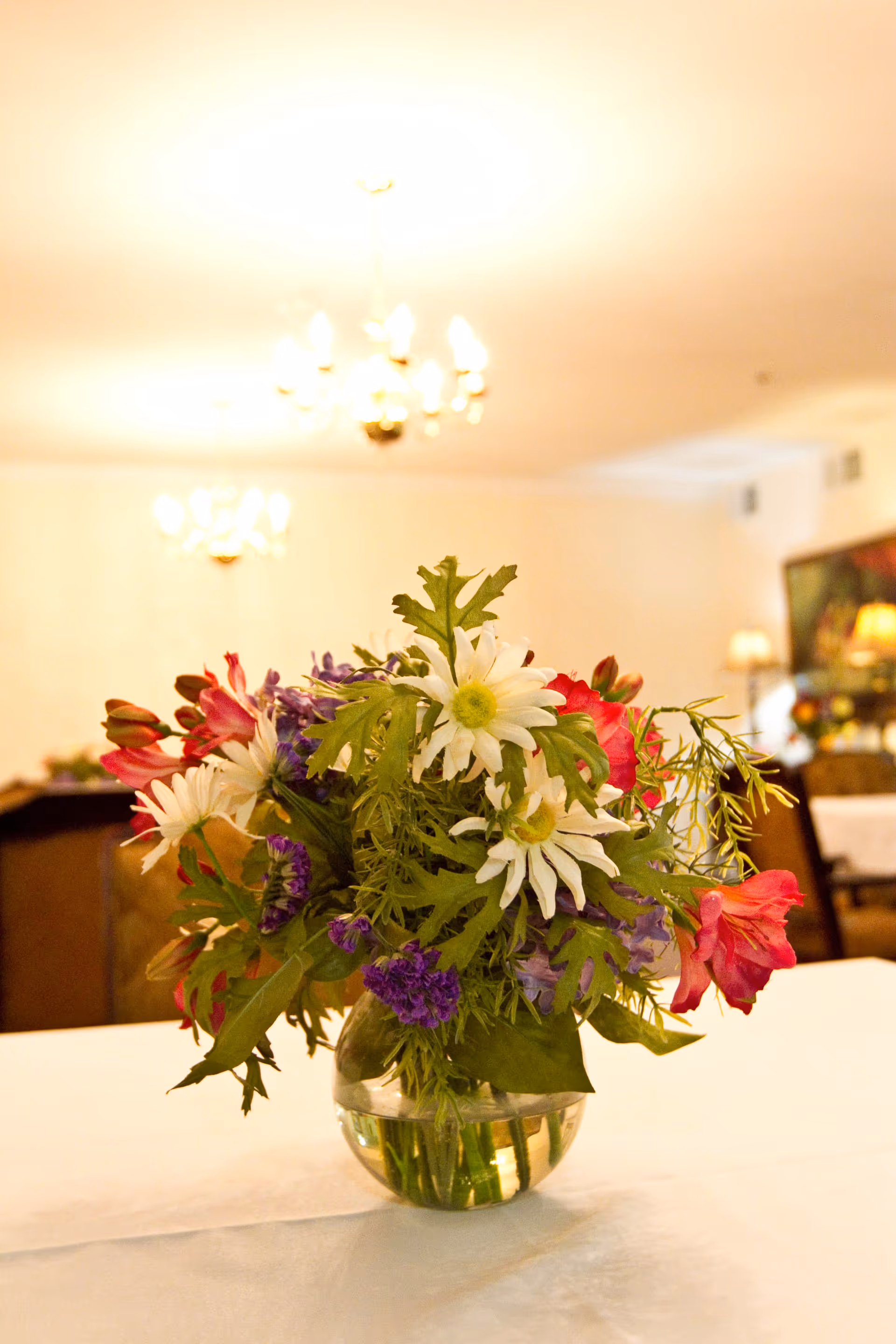 A colorful floral centerpiece in a glass vase sits on a table in a softly lit dining room with chandeliers.