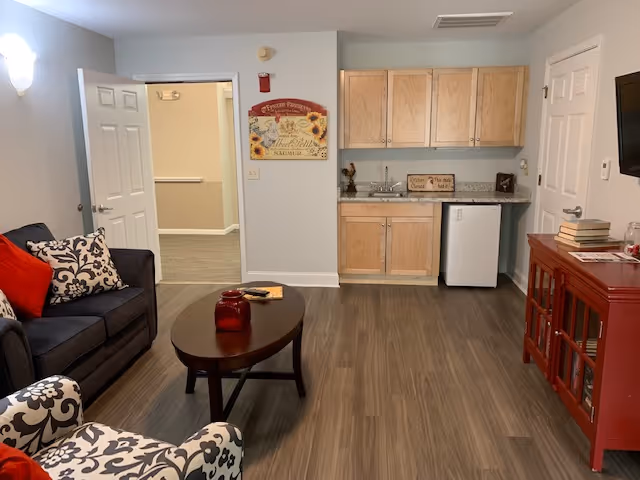 A cozy living area with a dark gray couch adorned with patterned and red pillows, a patterned armchair, and a wooden oval coffee table with red decorative items. The room has wood flooring and light-colored walls. On the right side, there is a small kitchenette with wooden cabinets, a granite countertop, a sink, and a mini refrigerator. A red wooden cabinet with books and decorative items is also visible. An open door leads to a hallway with carpeted flooring.