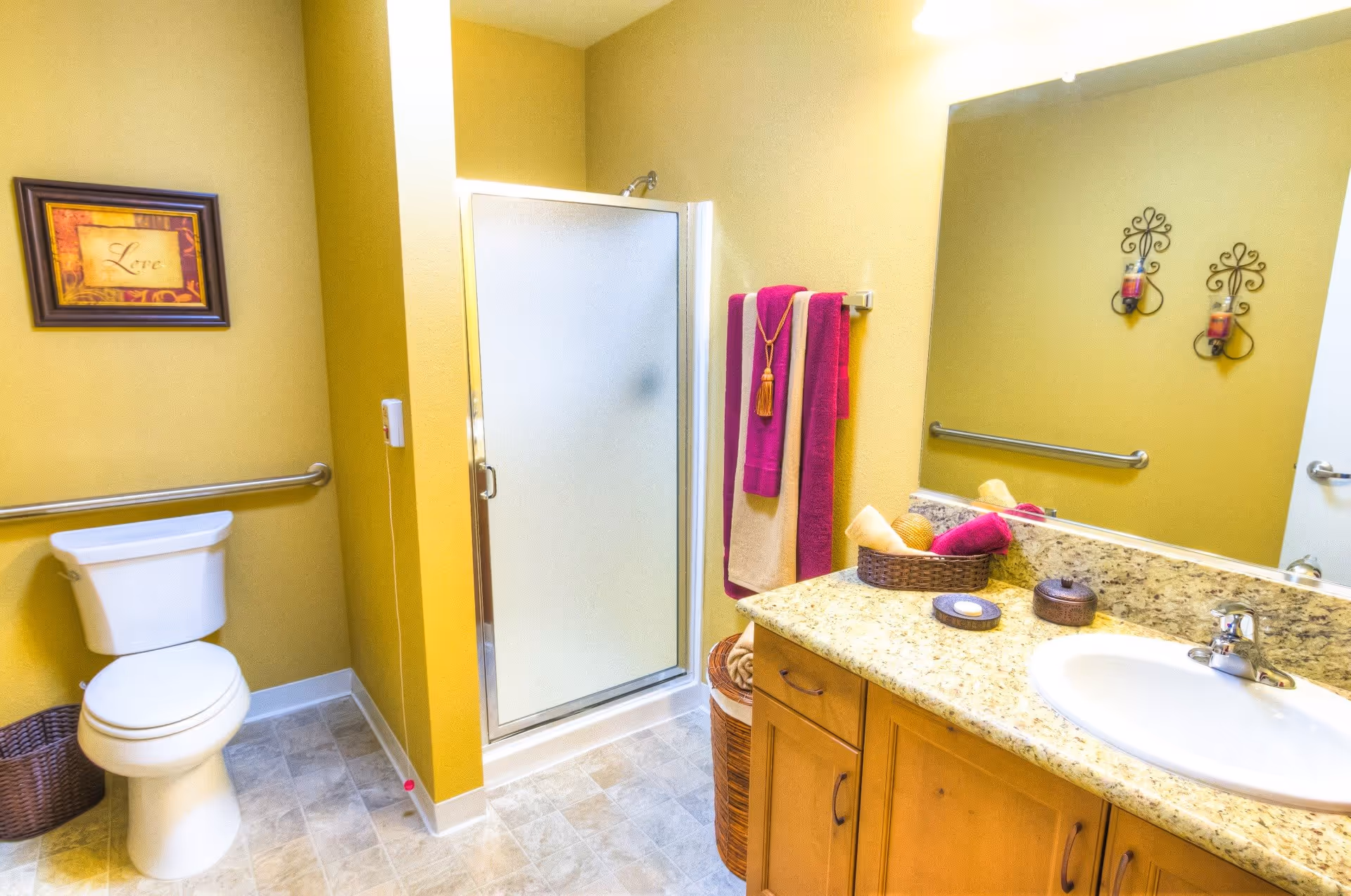 A clean and well-lit bathroom with yellow walls featuring a white toilet, a shower with a frosted glass door, a granite countertop with a sink, and wooden cabinets. There are colorful towels hanging on a rack and decorative wall art including a framed picture with the word 'Love' and two candle holders on the wall above the sink.
