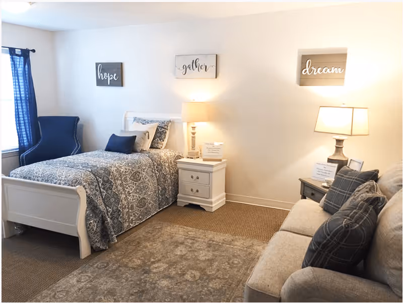 A cozy bedroom featuring a single bed with patterned bedding and pillows, a blue armchair near a window with blue curtains, two bedside tables each with a lamp, and a beige sofa with plaid pillows. The walls have three decorative signs with the words 'hope', 'gather', and 'dream'.