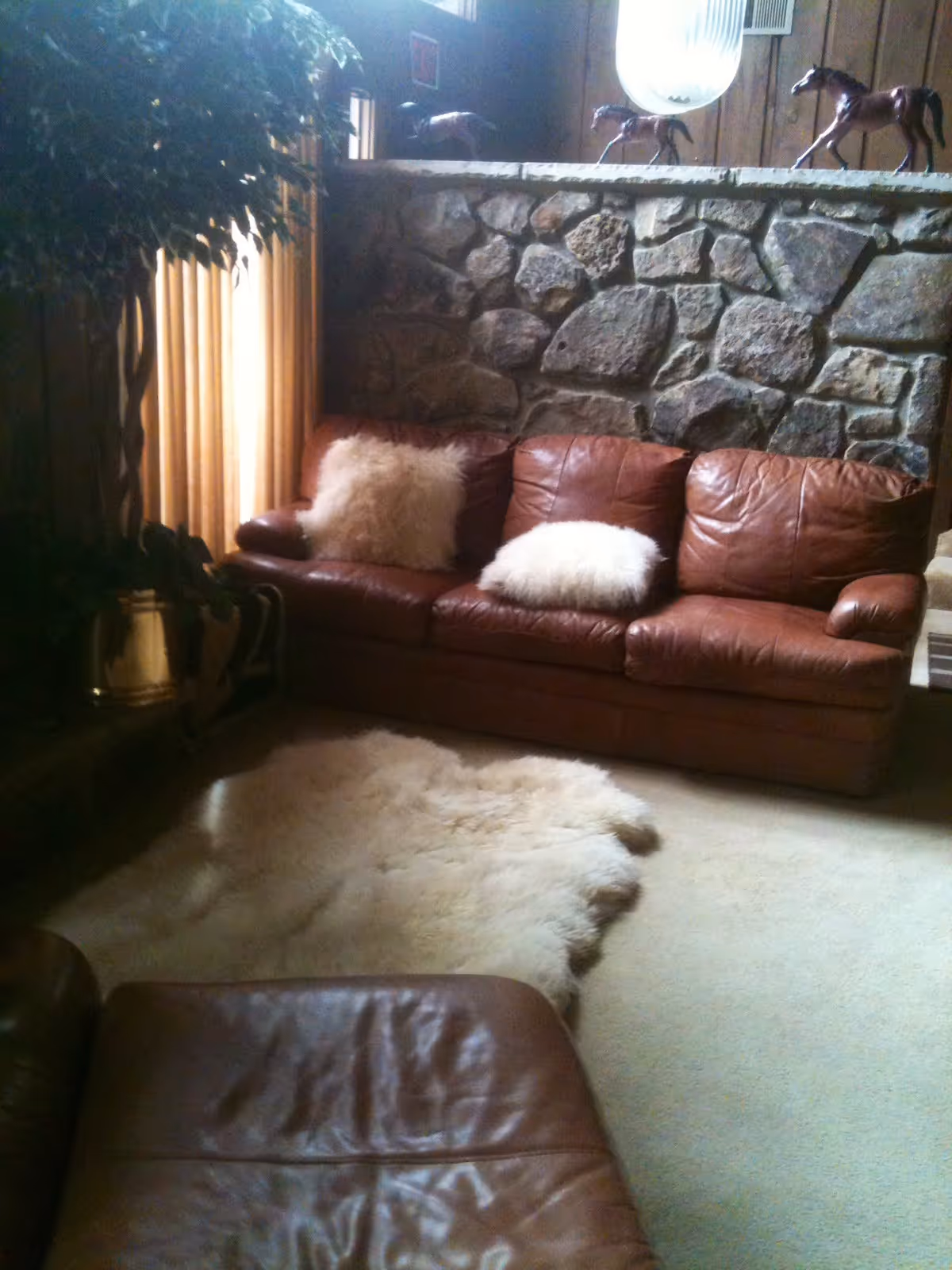 A cozy living room with a brown leather sofa adorned with two fluffy white pillows. There is a stone wall behind the sofa with three horse figurines placed on top. A large leafy plant is situated to the left near a window with beige curtains. A soft, light-colored rug lies on the carpeted floor in front of the sofa.