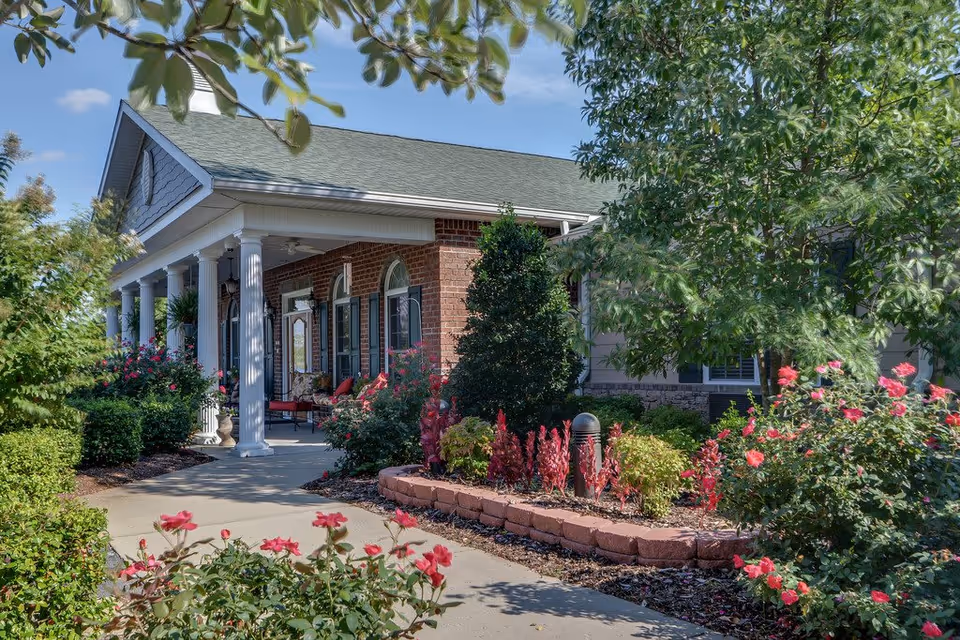 Exterior view of a brick building with white columns and a green roof, surrounded by a well-maintained garden with various shrubs, flowering plants, and trees under a clear blue sky.