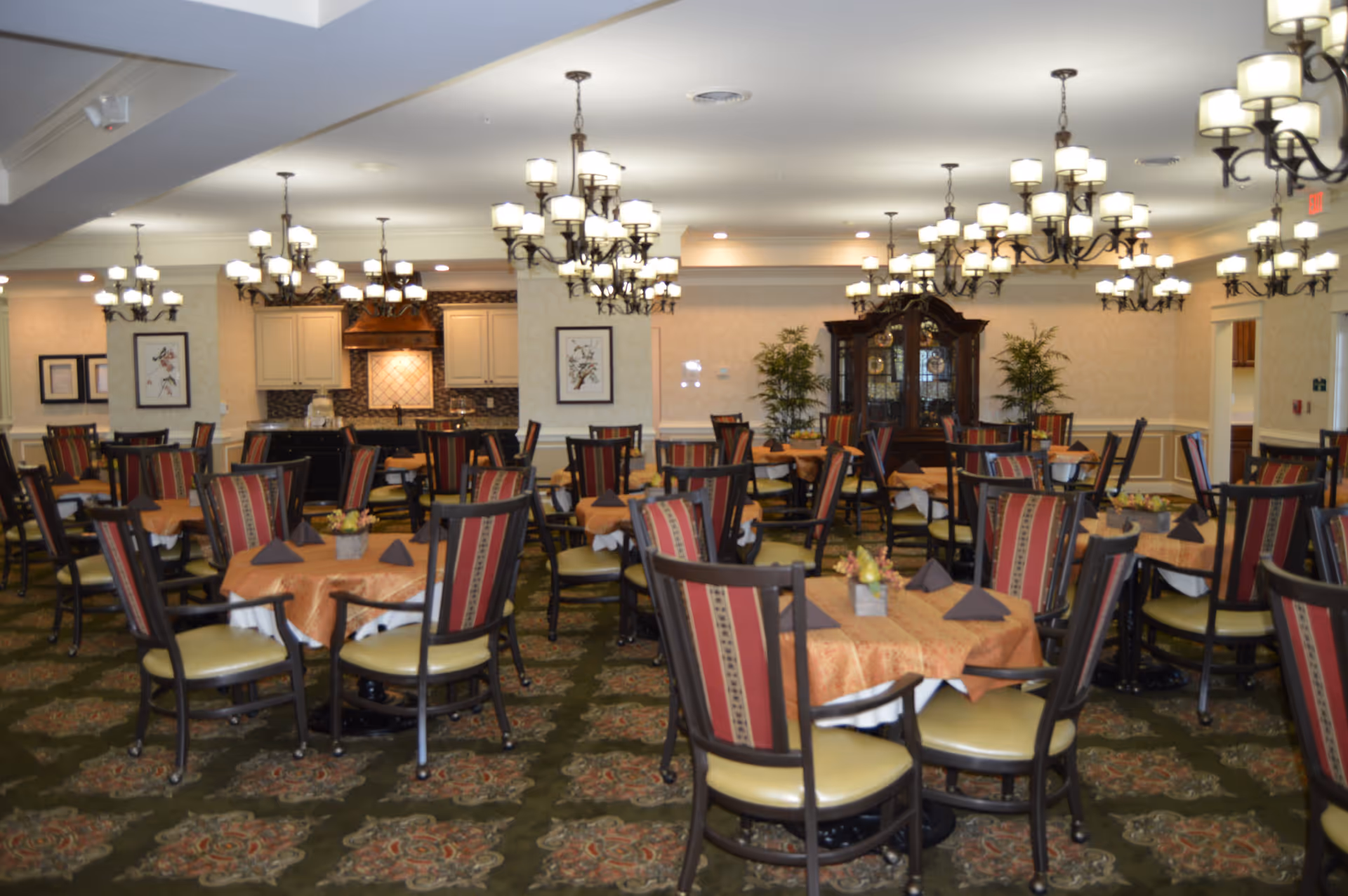Spacious dining room with multiple round tables and upholstered chairs under chandeliers and a buffet area in the back.