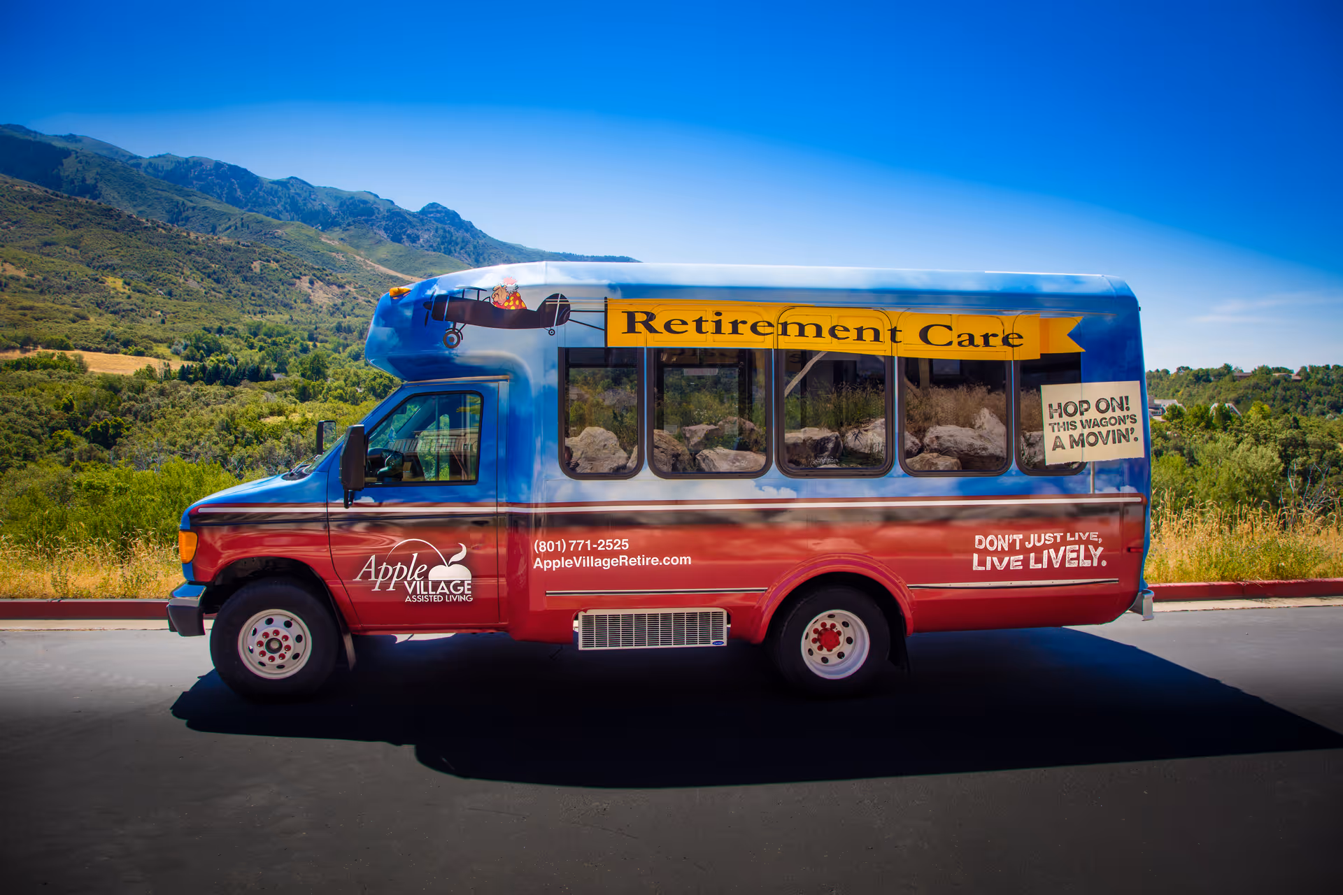 A colorful Apple Village retirement-care shuttle bus parked on a road with hills and a blue sky in the background.