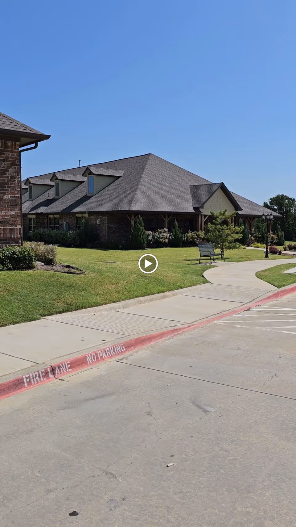 Exterior view of a single-story assisted living facility building with a large sloped roof, brick and siding walls, surrounded by a well-maintained lawn and landscaping. A curved sidewalk leads to the entrance, with a bench and a streetlamp nearby. The sky is clear and blue.