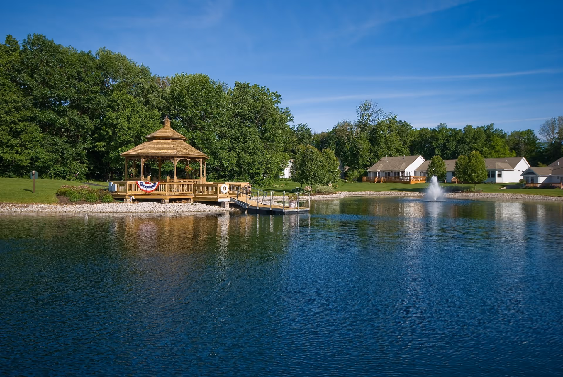 A peaceful outdoor scene featuring a wooden gazebo decorated with a patriotic banner, situated on the edge of a pond with a small dock. The pond has a fountain in the center, and several houses and trees are visible in the background under a clear blue sky.