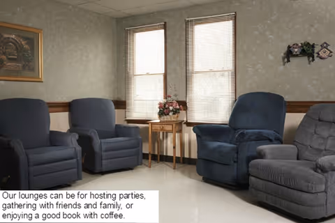 A cozy lounge area with four upholstered armchairs arranged around a small wooden side table holding a floral arrangement. Two large windows with blinds allow natural light into the room. The walls are decorated with a framed painting and a small wall shelf with decorative items.