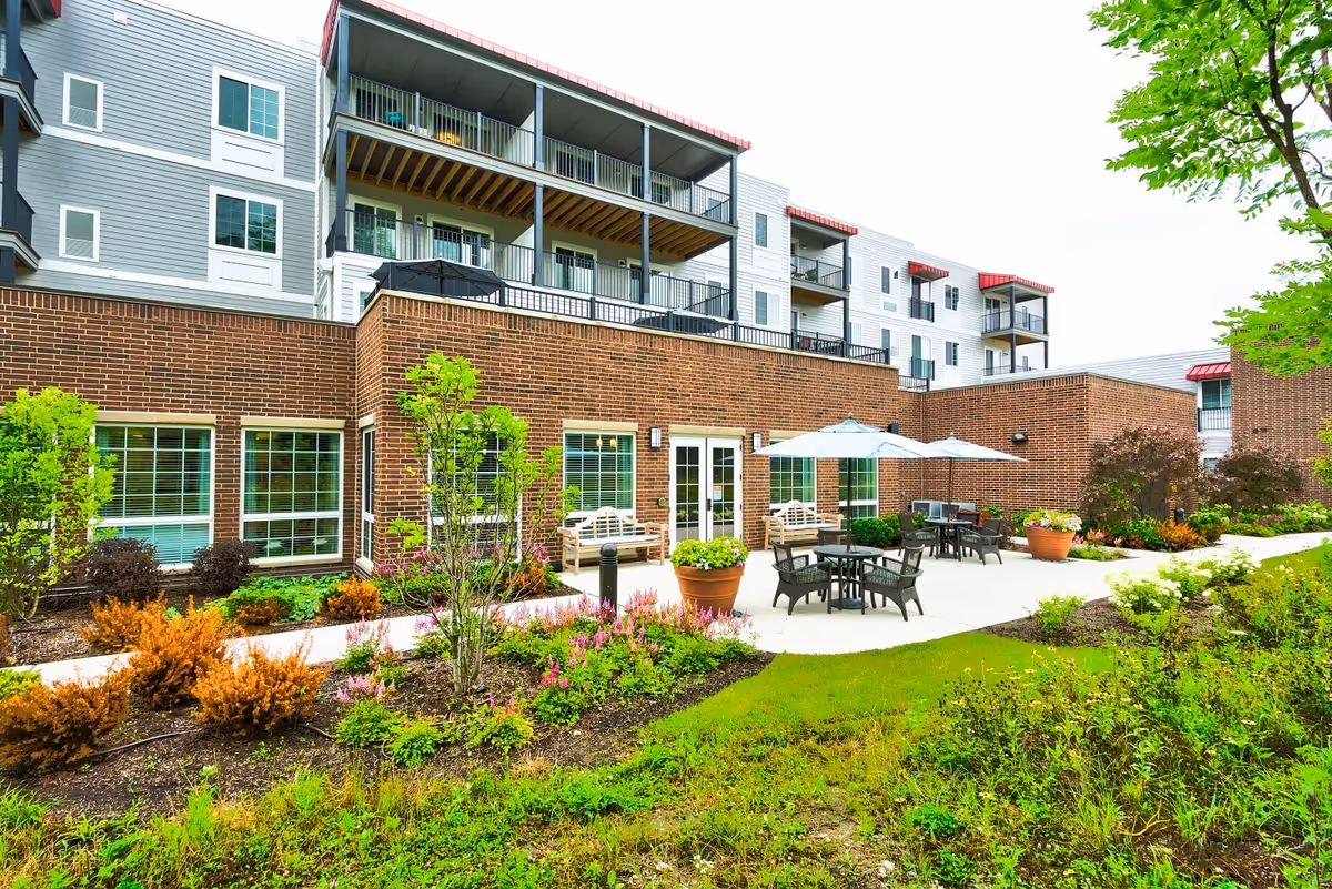 Outdoor patio area at Palos Heights Senior Living with tables, chairs, and umbrellas surrounded by landscaped garden beds and greenery, adjacent to a multi-story building with balconies and large windows.