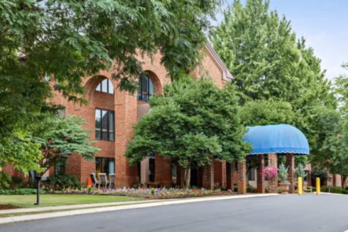 Brick-faced senior living building front with a blue-awned entrance, trees and landscaped driveway.