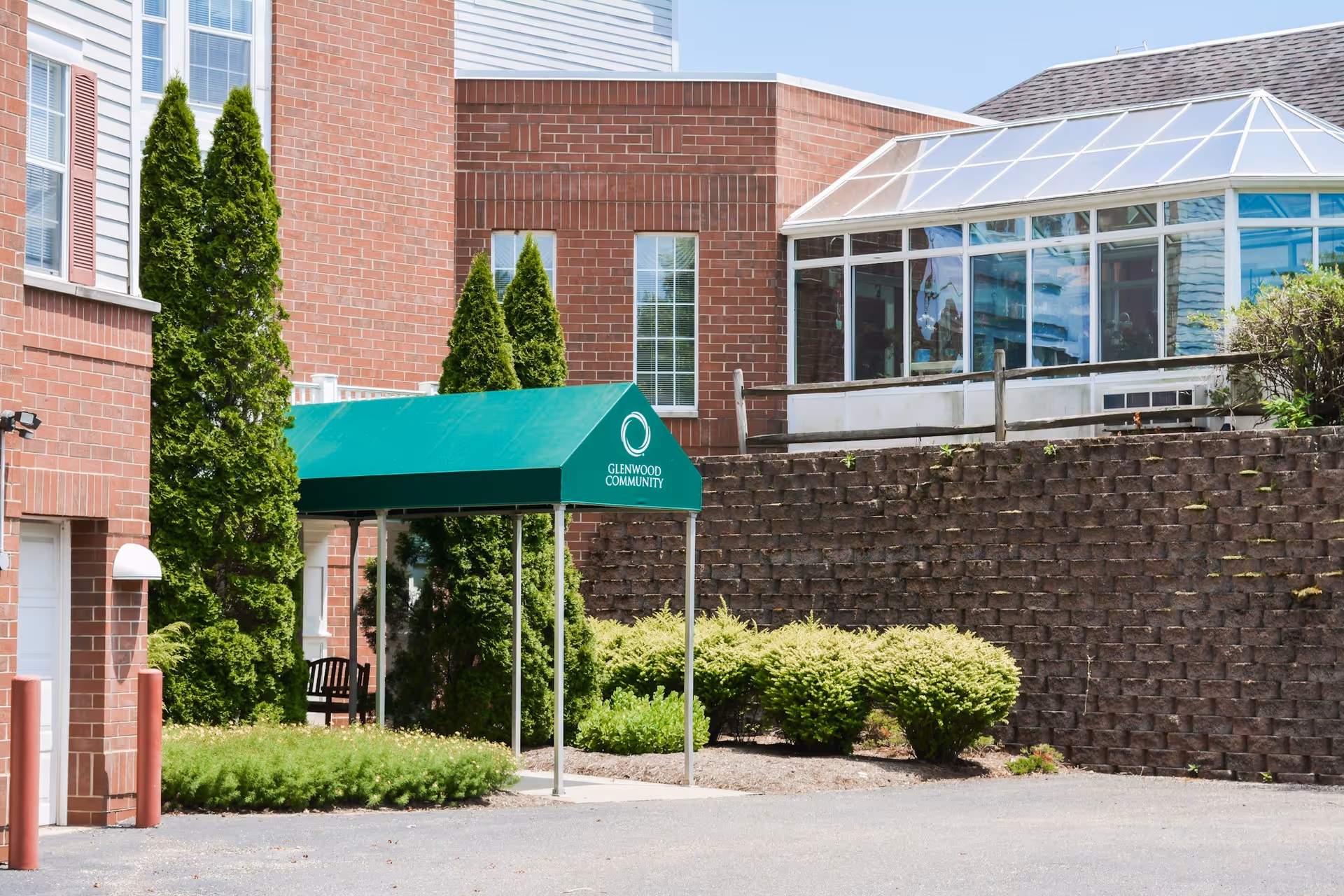 Exterior view of The Glenwood Community building showing a brick facade, green awning with the community name, tall evergreen trees, shrubs, and a glass sunroom on a sunny day.