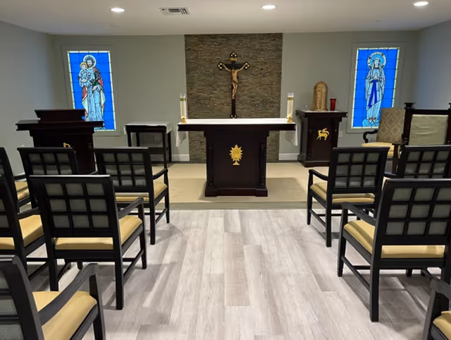 Interior view of a small chapel or prayer room with rows of chairs facing an altar. The altar has a crucifix mounted on a stone wall behind it, flanked by two stained glass windows depicting religious figures. There are candles and religious symbols on the altar and surrounding furniture.