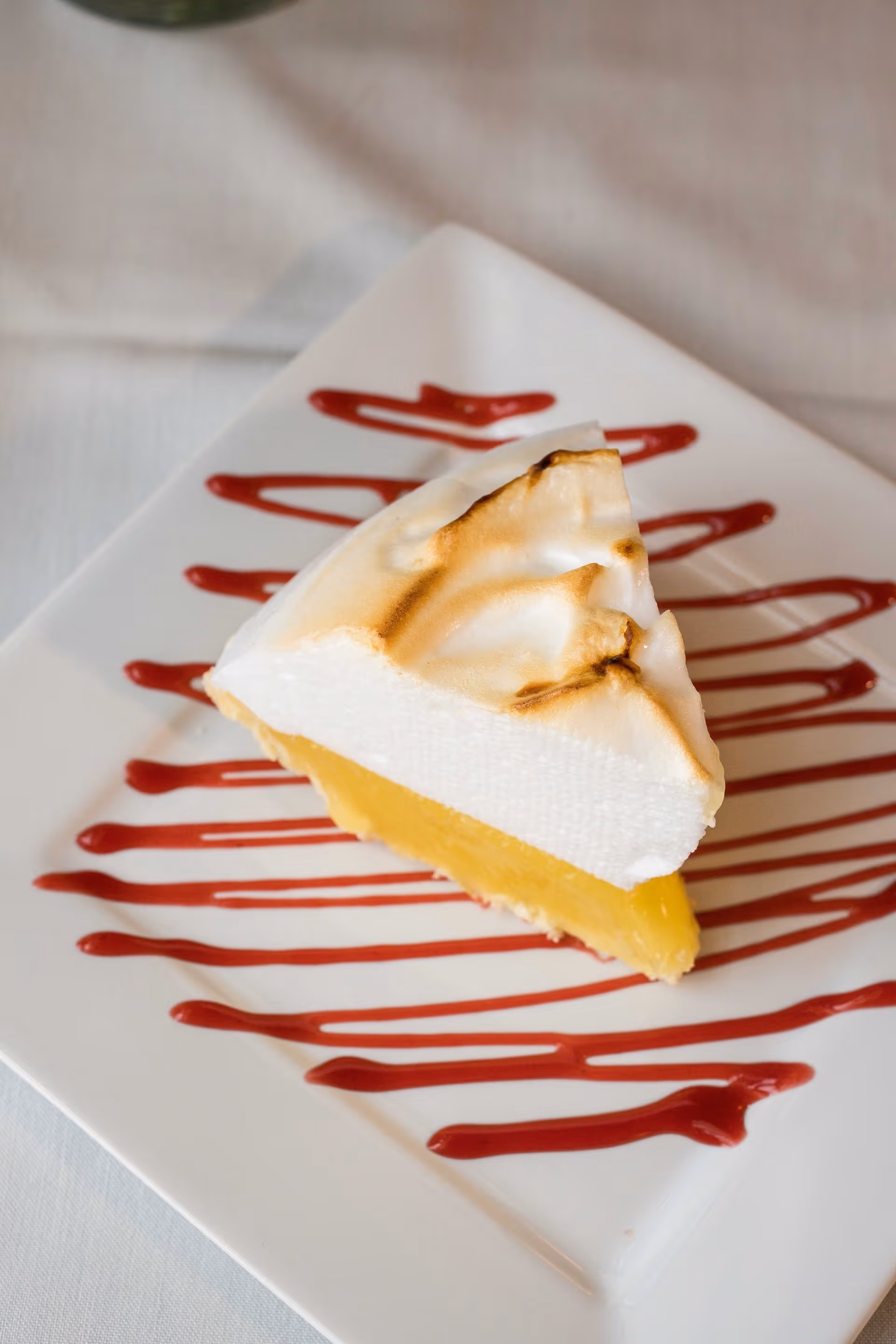 A slice of lemon meringue pie on a white square plate with red sauce drizzled in a zigzag pattern underneath the pie.