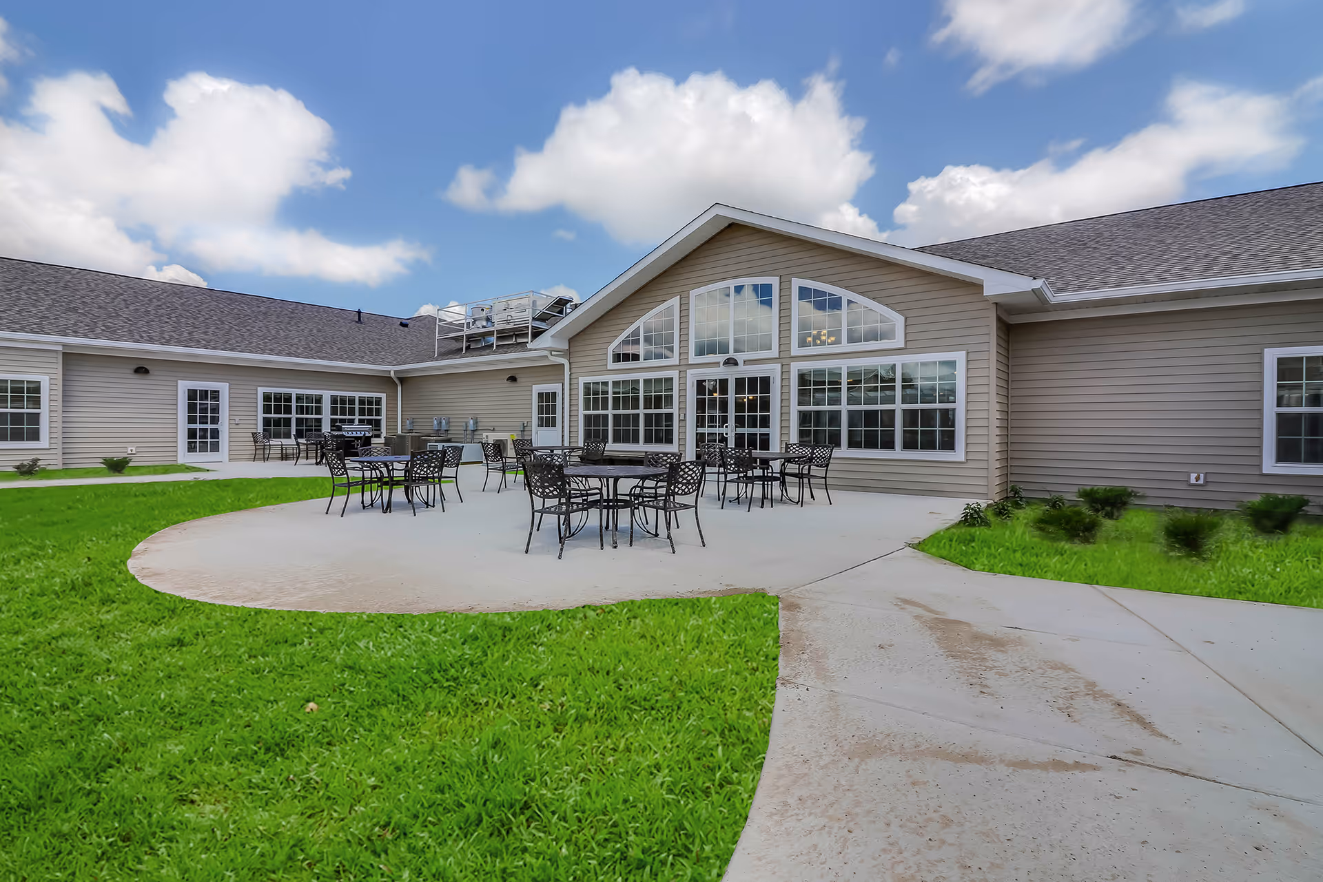 Outdoor patio area of a senior living facility with several black metal tables and chairs arranged on a concrete circular patio. The building has beige siding, multiple large windows, and a peaked roof. The patio is surrounded by green grass and a concrete walkway. The sky is blue with scattered clouds.