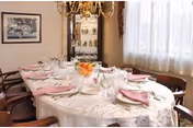 A dining room with a large oval table covered with a white lace tablecloth, set with plates, glasses, and pink folded napkins. There is a chandelier hanging above the table, a glass cabinet with decorative items in the background, and a window with sheer white curtains letting in natural light.