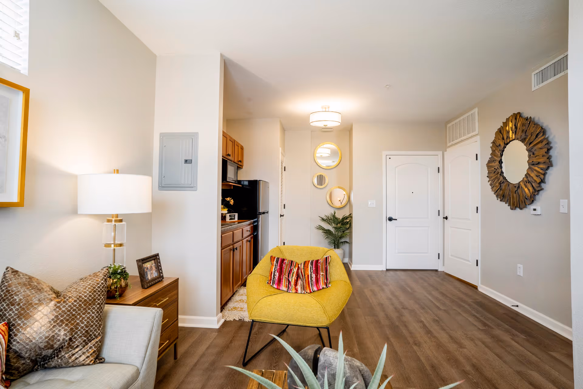 A bright and modern living area with a yellow armchair featuring a colorful cushion, a beige sofa with patterned pillows, a wooden side table with a lamp and framed photo, and a small kitchen area with wooden cabinets and stainless steel appliances. The room has light-colored walls, wood flooring, decorative round mirrors on the wall, and a door leading to another room.