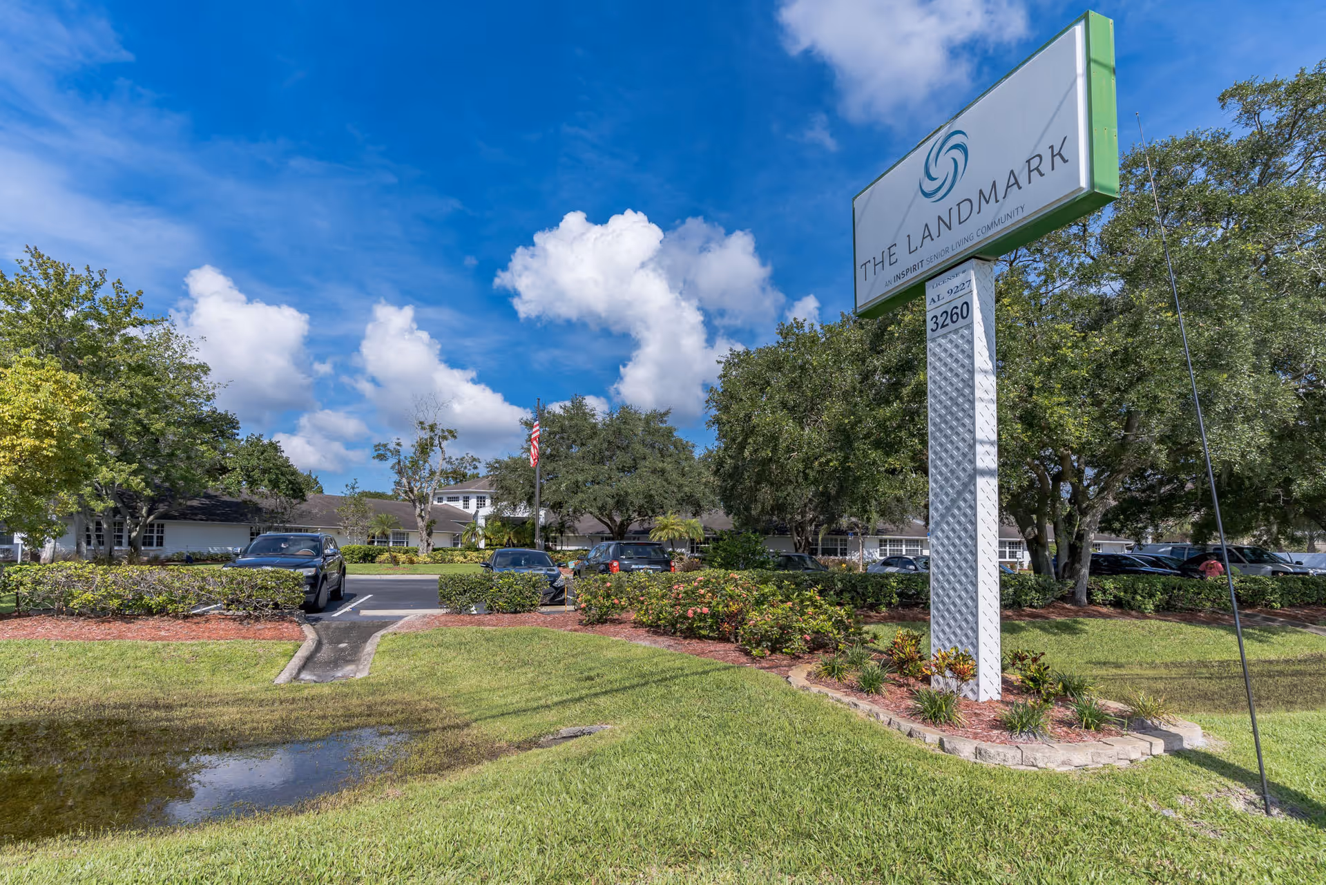 Outdoor view of The Landmark senior living community sign with a parking lot, trees, and a partly cloudy blue sky in the background.