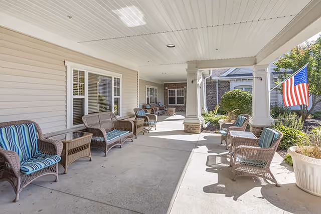 Covered outdoor patio area with wicker chairs and small tables arranged along the sides. The patio is attached to a building with beige siding and white trim. An American flag is displayed on a pole near the edge of the patio, and there are plants and bushes in the surrounding garden area.