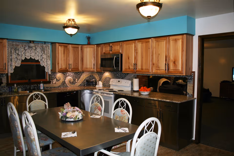 A kitchen and dining area with wooden cabinets, a stove, microwave, dishwasher, and a sink under a window with lace curtains. A rectangular dining table with six chairs, each with floral upholstery, is set with napkins and silverware. A decorative centerpiece is on the table, and a bowl of oranges is on the kitchen counter. The room is warmly lit with ceiling lights.