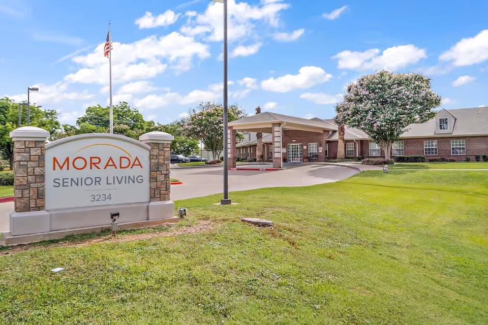 Exterior view of Morada Senior Living facility with a large sign in the foreground displaying the name and address. The building is a single-story brick structure with a covered entrance, surrounded by green grass, trees, and a clear blue sky with some clouds.