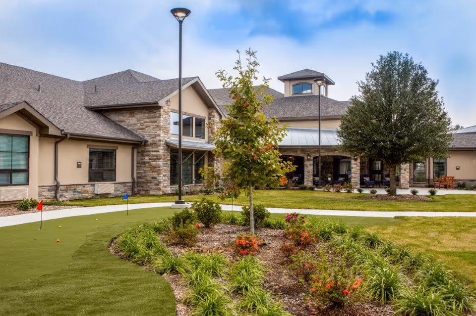 Exterior front of a senior living building with a landscaped courtyard, small putting green, trees, and a covered entrance.