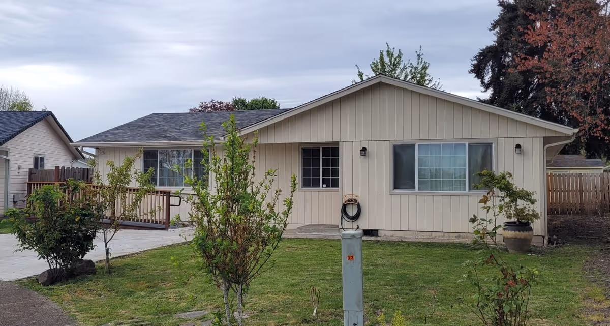 Single-story beige residential building with a sloped roof, two windows, a door, and a small front yard with grass, bushes, and a concrete driveway. The sky is cloudy.