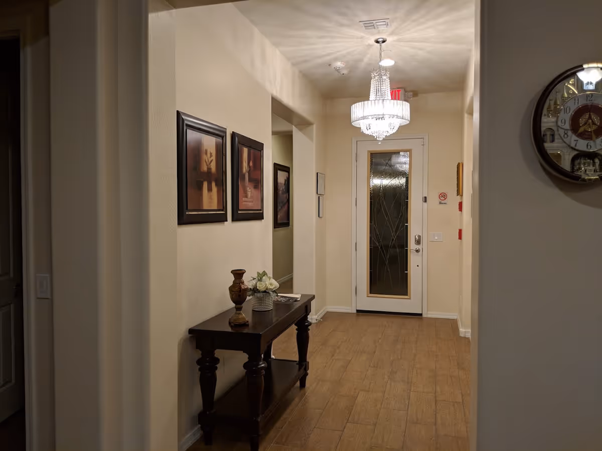 A hallway with beige walls and wooden flooring, featuring a dark wooden console table with decorative items including a vase and flowers. Three framed artworks hang on the left wall, and a decorative glass door is at the end of the hallway. A chandelier light fixture hangs from the ceiling, and a round wall clock is visible on the right wall.