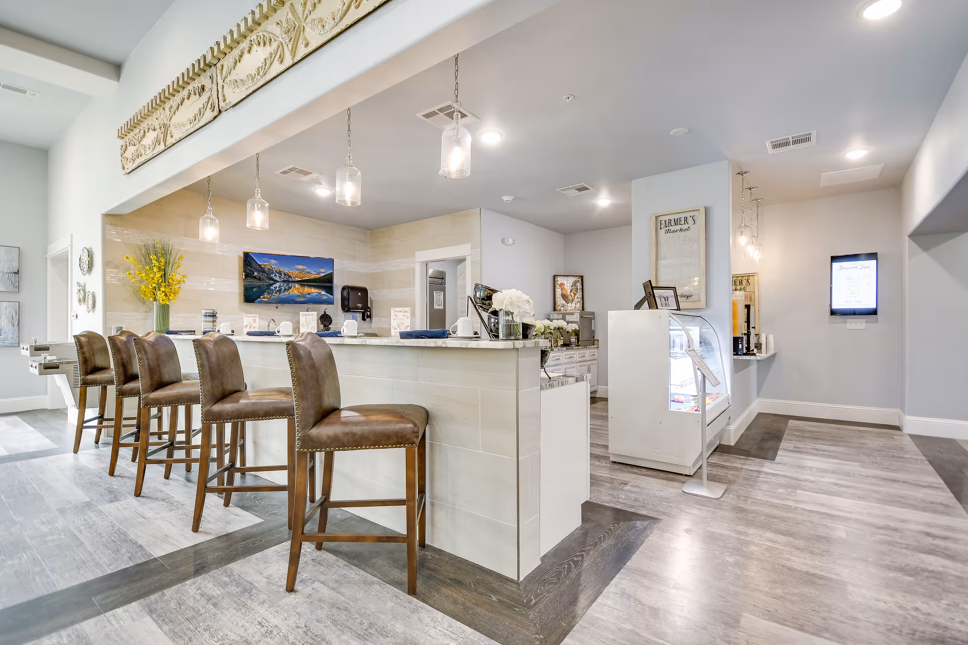 Bright modern common-area café with a long counter, leather barstools, pendant lights, and a small display case.