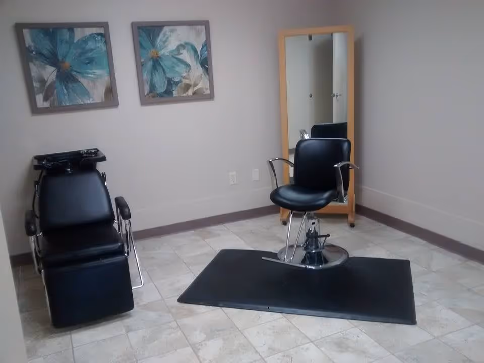 A small salon area with a black salon chair on a black mat in front of a full-length mirror with a wooden frame. To the left, there is a black shampoo chair with a sink attached. Two framed paintings of blue flowers hang on the light-colored wall.