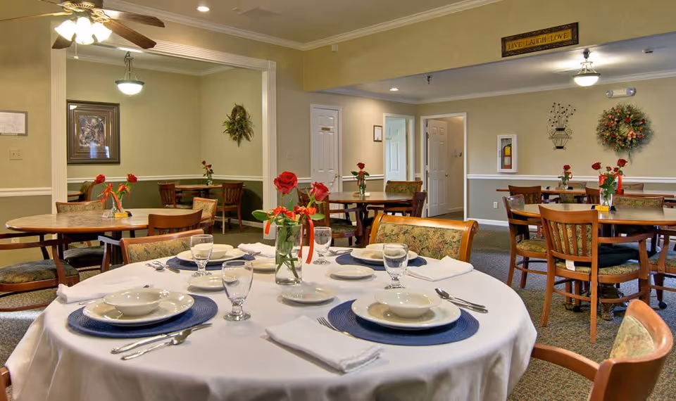 A dining room in a senior living facility with several round tables set for meals. The closest table is covered with a white tablecloth and set with plates, bowls, glasses, and silverware. Each table has a vase with red roses tied with red ribbons. The room has beige walls decorated with framed pictures and wreaths, and ceiling fans with lights.