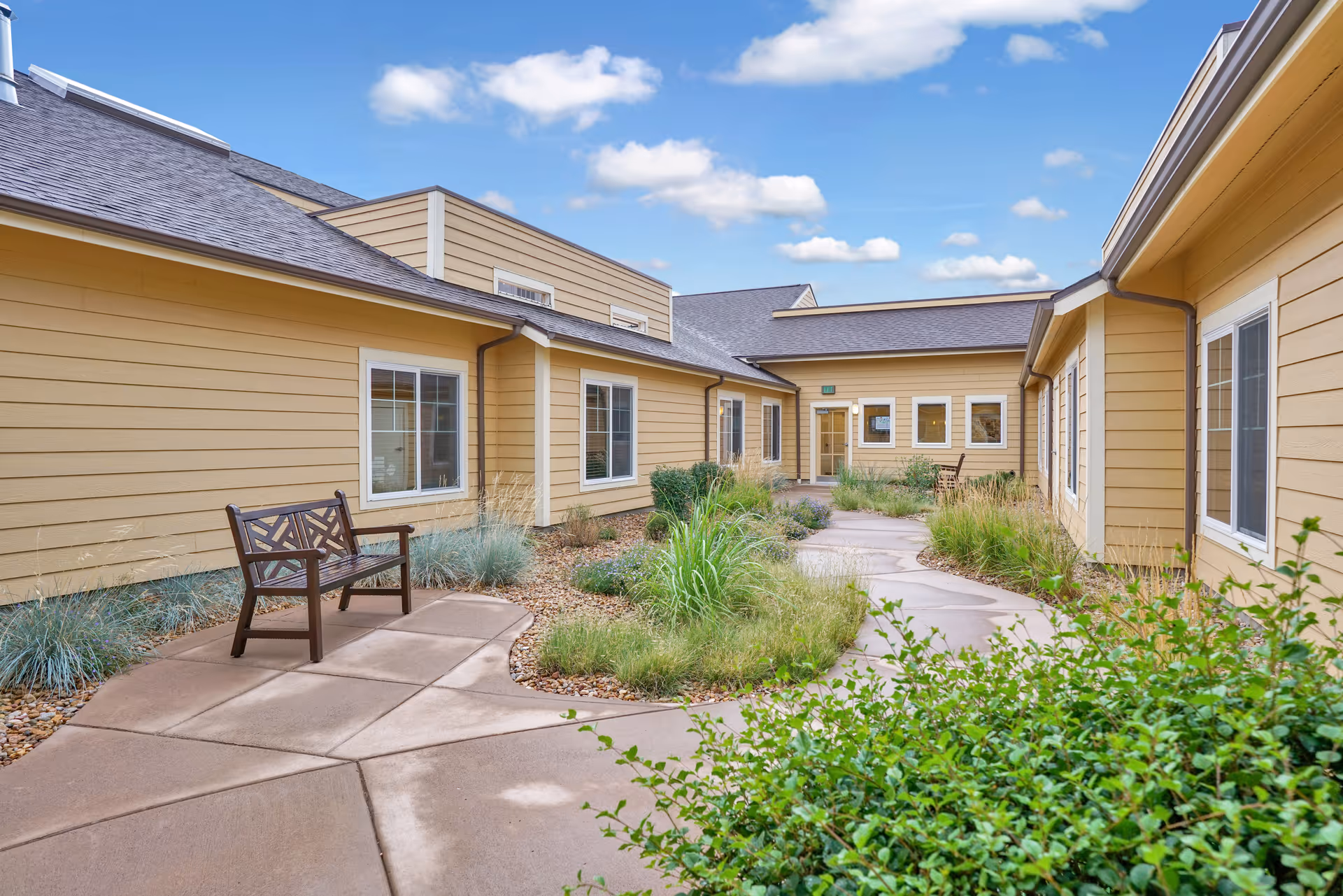 Outdoor courtyard area at Seven Lakes Memory Care with a paved walkway, green shrubs, ornamental grasses, and two wooden benches against the beige exterior walls of the building under a partly cloudy blue sky.