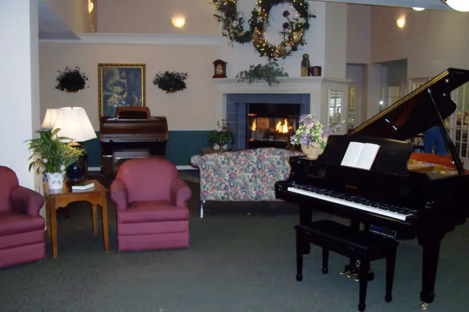 A cozy living room area with a black grand piano and an open sheet music book on it. There are two red armchairs with a wooden side table and a lamp between them on the left. In the background, a floral-patterned sofa faces a lit fireplace with a decorative wreath above it. A wooden upright piano and framed artwork are also visible on the back wall, along with some hanging plants.