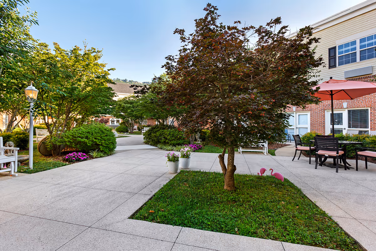 Outdoor courtyard area with paved walkways, green grass, and various trees and bushes. There are white benches, potted flowers, and a patio table with chairs and a red umbrella. The building exterior is visible in the background under a clear blue sky.
