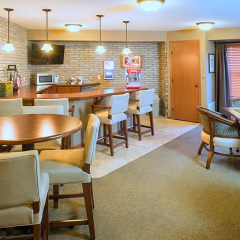 A cozy common area in a senior living facility featuring a round wooden table with upholstered chairs, a bar counter with high chairs, a microwave, a popcorn machine, and a wall-mounted TV. The room has a brick accent wall, warm lighting, and a door leading to another room.