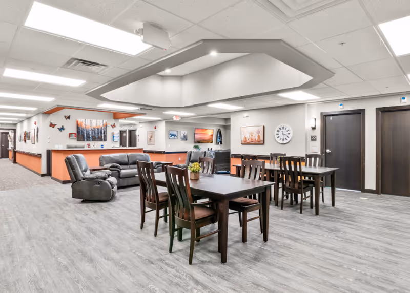 A spacious and well-lit common area in a senior living facility featuring multiple dark wood tables with chairs, a comfortable gray sofa and recliner, wall art including paintings and butterfly decorations, a large clock, and doors leading to other rooms.