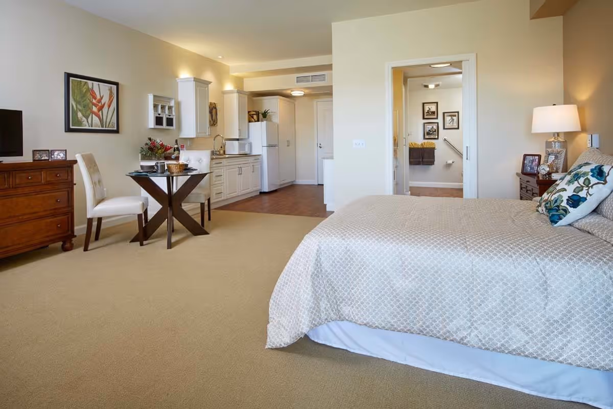 A spacious senior living studio apartment featuring a bed with patterned bedding, a bedside table with a lamp and framed photos, a small dining table with two chairs, a wooden dresser with a TV and picture frames, and a kitchenette with white cabinets, a refrigerator, microwave, and sink. The room has beige walls and carpet, with a doorway leading to a bathroom visible in the background.