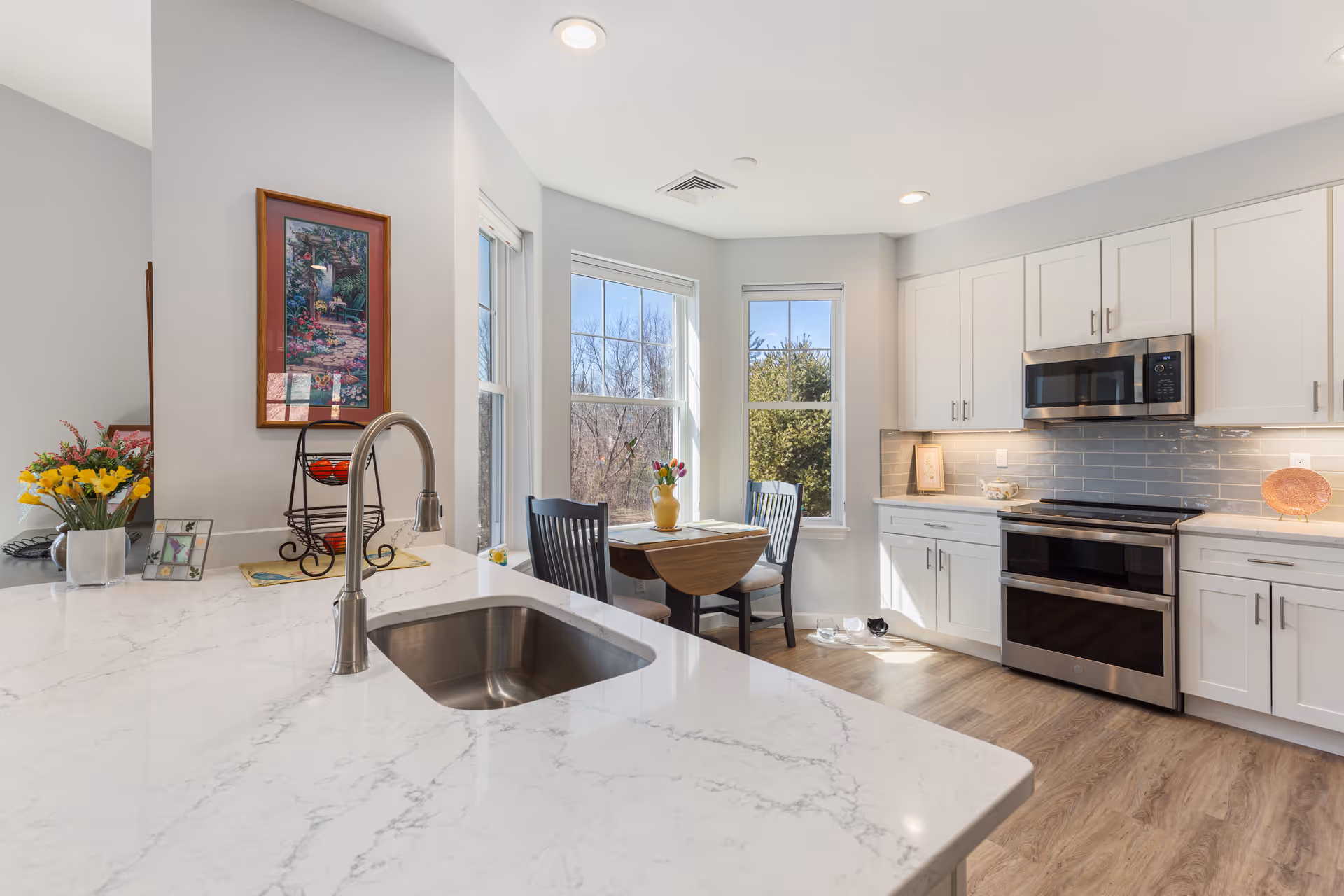Bright modern kitchen with a marble island and sink, stainless steel appliances, white cabinets, and a small dining table by bay windows.