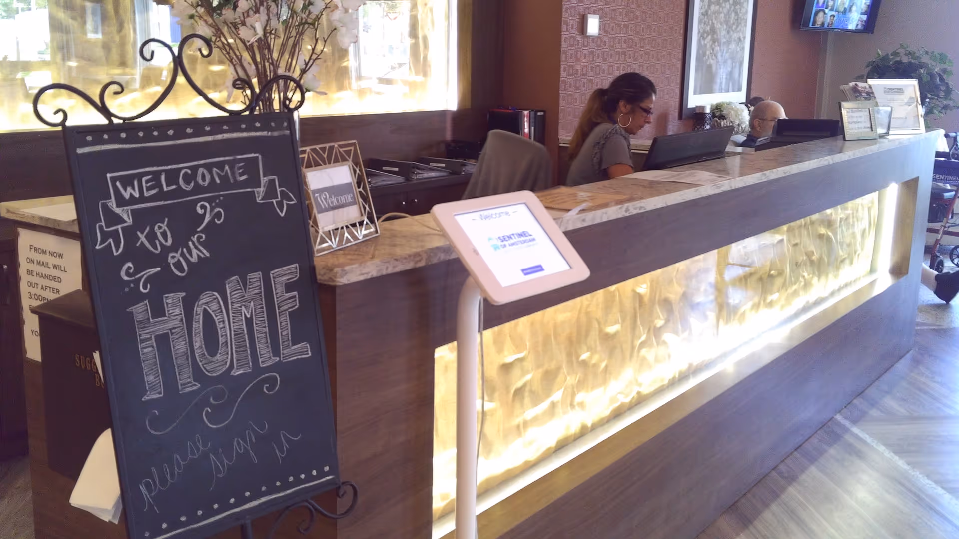 Reception desk area at The Sentinel of Amsterdam with two staff members working behind the counter. A decorative chalkboard sign reads 'Welcome to our Home, please sign in.' The desk features a lighted panel and a tablet for visitor sign-in. There are flowers and framed welcome signs on the counter.