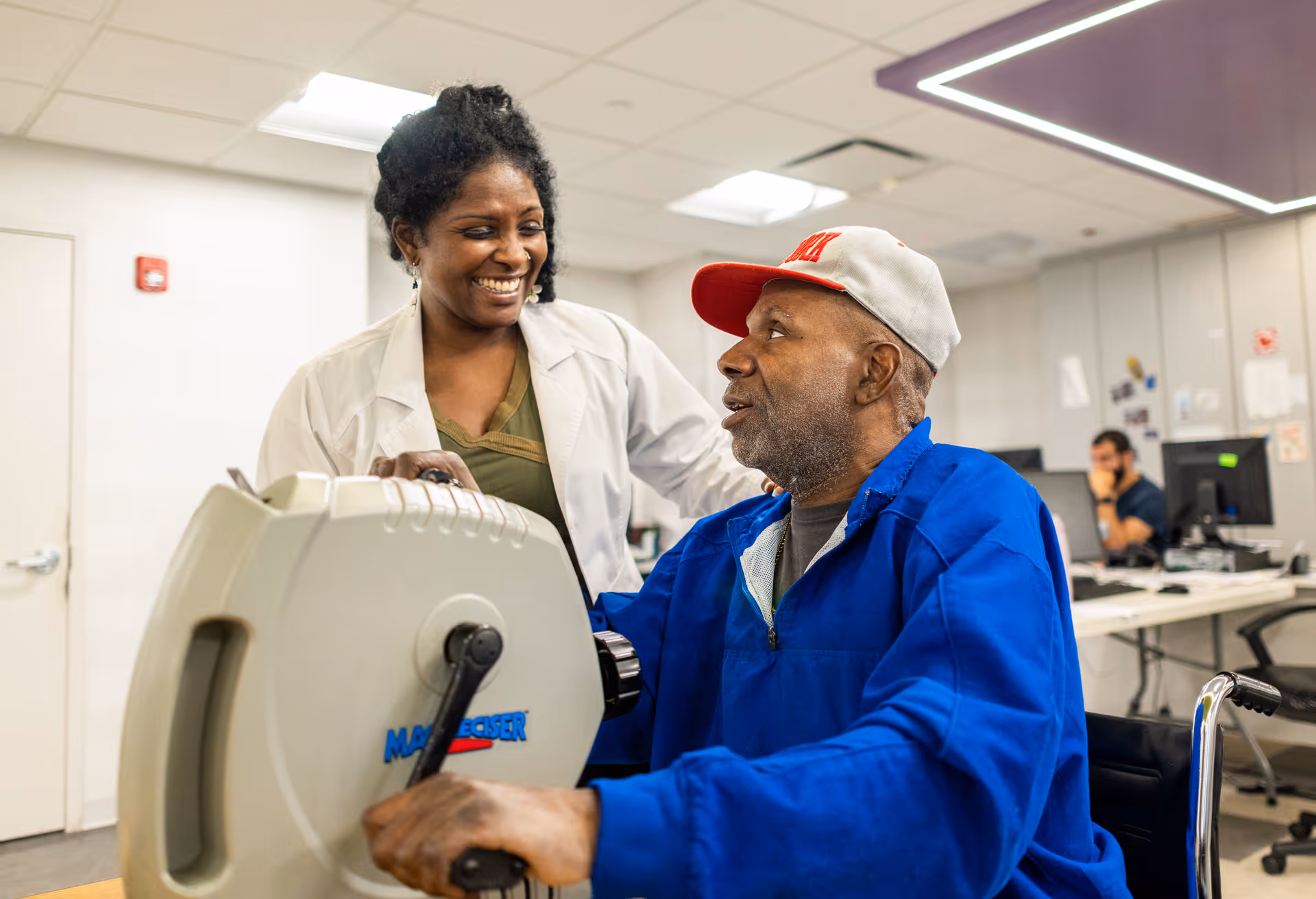 A healthcare professional in a white coat smiles and interacts with an elderly man in a blue jacket and gray cap who is using a hand exercise machine in a clinical setting with desks and computers in the background.