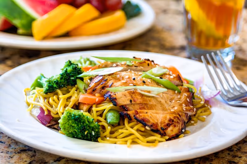 A plate of grilled chicken served on a bed of yellow noodles with broccoli, carrots, and green onions, garnished with sesame seeds. In the background, there is a blurred plate of fresh fruit and a glass of iced tea with lemon.