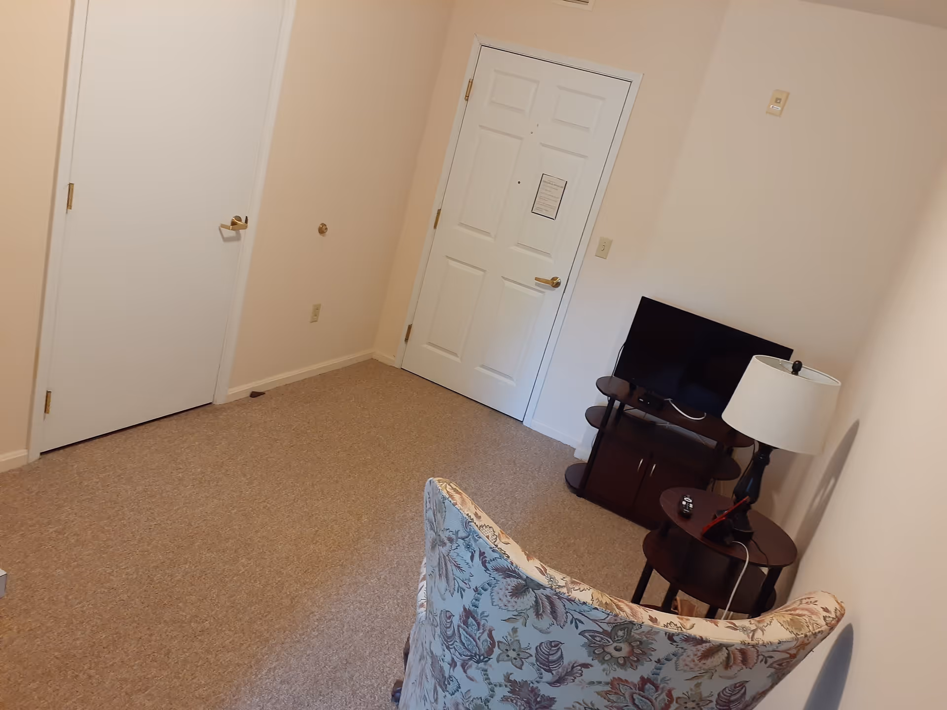 A small room with beige walls and carpeted floor featuring a floral-patterned armchair, a small wooden side table with a lamp and remote controls, and a flat-screen TV on a dark wooden TV stand. There are two white doors, one likely an entrance door and the other possibly a closet or bathroom door.