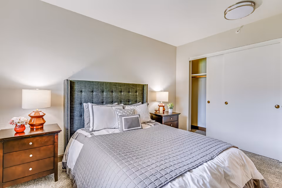 A neatly made bed with a gray quilted bedspread and multiple pillows in a bedroom. The bed has a dark upholstered headboard. On either side of the bed are wooden nightstands with table lamps and small decorative items. There is a closet with sliding white doors on the right side of the room. The walls are painted light beige and the floor is carpeted.