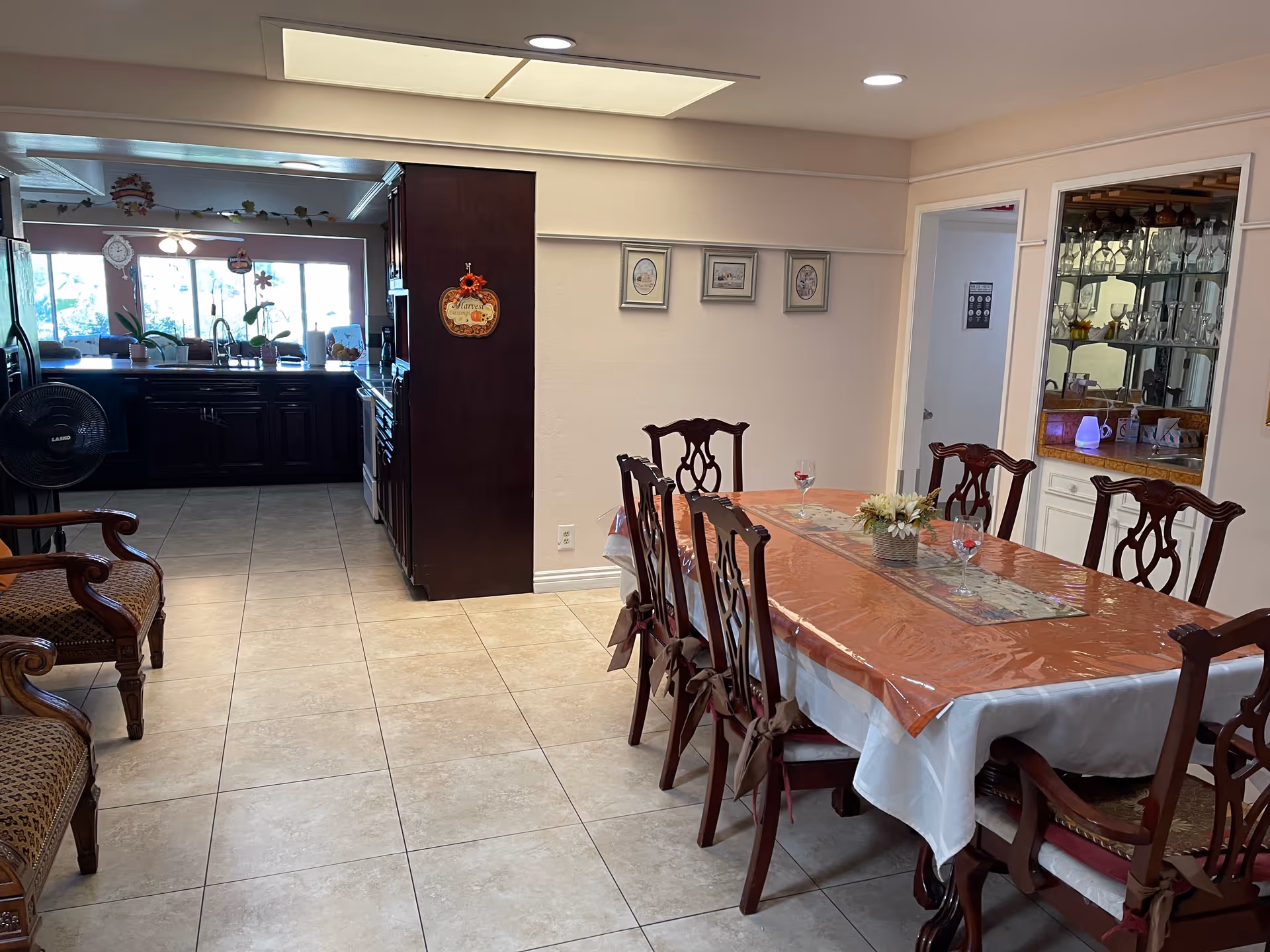 A dining area with a long wooden table covered with a plastic tablecloth and a floral centerpiece. There are eight wooden chairs around the table. To the left, there are two upholstered armchairs. In the background, there is a kitchen with dark wooden cabinets, a sink, and large windows letting in natural light. On the right wall, there is a built-in glass cabinet displaying glassware.